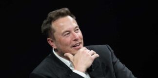 Man in black suit smiling and touching his face.