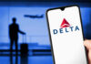 Delta STUNS Congress – No More VIP Perks! Person holding smartphone displaying Delta logo airport background