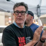 A man in a black t-shirt with a writer's guild logo, clapping at a public event
