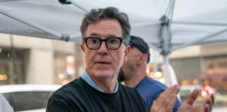 A man in a black t-shirt with a writer's guild logo, clapping at a public event