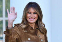 Woman waving and smiling in brown coat.