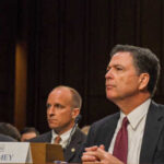 Government officials seated during a hearing with a nameplate in front of one