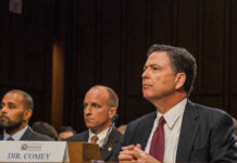 Government officials seated during a hearing with a nameplate in front of one