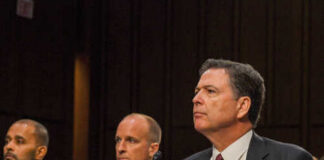 Government officials seated during a hearing with a nameplate in front of one
