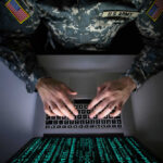 A soldier in U.S. Army uniform typing on a laptop with green code displayed on the screen