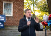 A man speaking at an outdoor event with campaign signs in the background