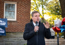 A man speaking at an outdoor event with campaign signs in the background