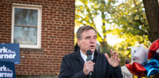 A man speaking at an outdoor event with campaign signs in the background