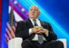 Man in suit sitting on stage with American flag.