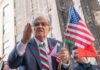 A man passionately speaking at a rally while holding American flags