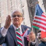 A man passionately speaking at a rally while holding American flags