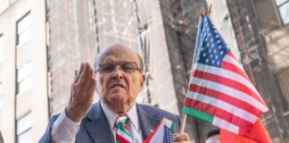 A man passionately speaking at a rally while holding American flags