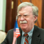 Classified Chaos: Bolton Deal Stuns D.C. Man with glasses smiling near small American flag