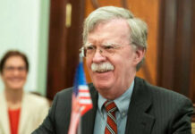 Classified Chaos: Bolton Deal Stuns D.C. Man with glasses smiling near small American flag