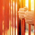 Hands gripping orange prison bars with sunlight in the background