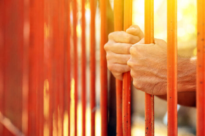 Male,Hands,Behind,Prison,Yard,Bars,,Incarcerated,Captivated,Person,In Hands gripping orange prison bars with sunlight in the background
