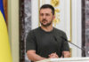 Zelensky Fires Reformer Amid War Tensions Man speaking at podium with microphones, Ukrainian flag behind.