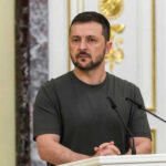 Zelensky Fires Reformer Amid War Tensions Man speaking at podium with microphones, Ukrainian flag behind.