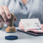 New Visa Rules Escalate U.S.–China Media Fight A hand holding a wooden stamp above a passport on a table