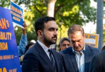 Dangerous Heat Pushes New York Into Action Man in suit speaking at outdoor press event with signs in background