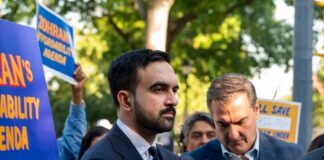 Man in suit speaking at outdoor press event with signs in background
