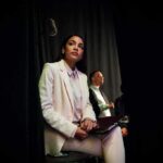 AOC’s “Crazy” Woke Era Comes Back Around Woman in a white suit sitting backstage with a folder