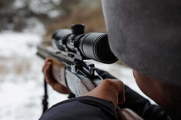 person aiming a rifle with a scope outdoors