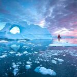 Greenland Control Deal Ignites Questions Iceberg with arch and red-sailed boat at colorful sunset