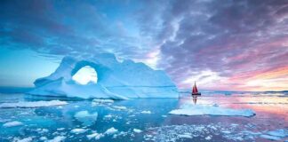 Iceberg with arch and red-sailed boat at colorful sunset