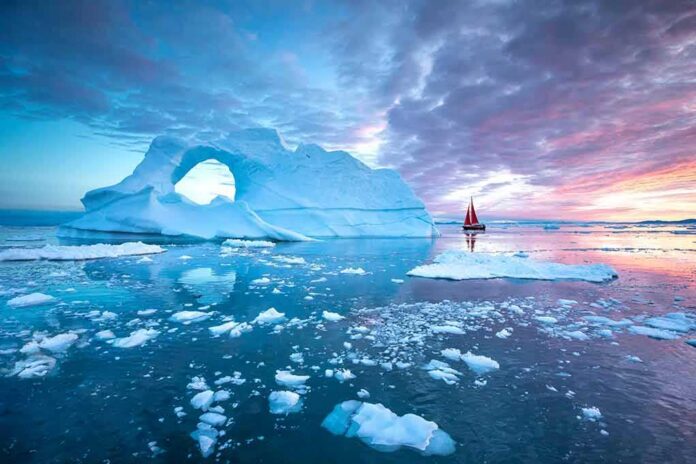 Iceberg with arch and red-sailed boat at colorful sunset