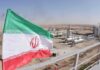 Iran Took The Hits—Where’s The Finish Line? Iran flag over an oil and gas facility in a desert landscape