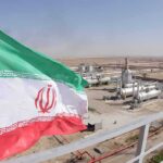 Iran Took The Hits—Where’s The Finish Line? Iran flag over an oil and gas facility in a desert landscape