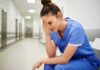 Debt-Strapped Hospitals Slash Wages, Stall Pay Nurse in blue scrubs sitting with head in hand