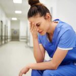 Debt-Strapped Hospitals Slash Wages, Stall Pay Nurse in blue scrubs sitting with head in hand
