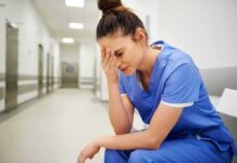 Debt-Strapped Hospitals Slash Wages, Stall Pay Nurse in blue scrubs sitting with head in hand