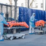 Healthcare workers in PPE move covered stretchers outside a facility