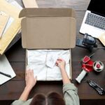 Person packing a cardboard box on a cluttered desk
