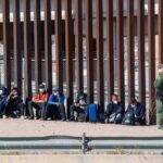 Group of people sitting by a tall border fence with an officer standing nearby